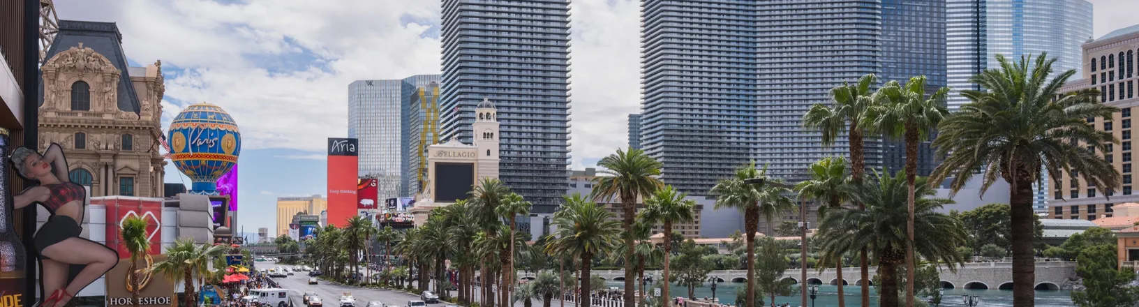 The Las Vegas Strip on a bright day, lined with palm trees and resort towers, with traffic and pedestrians.