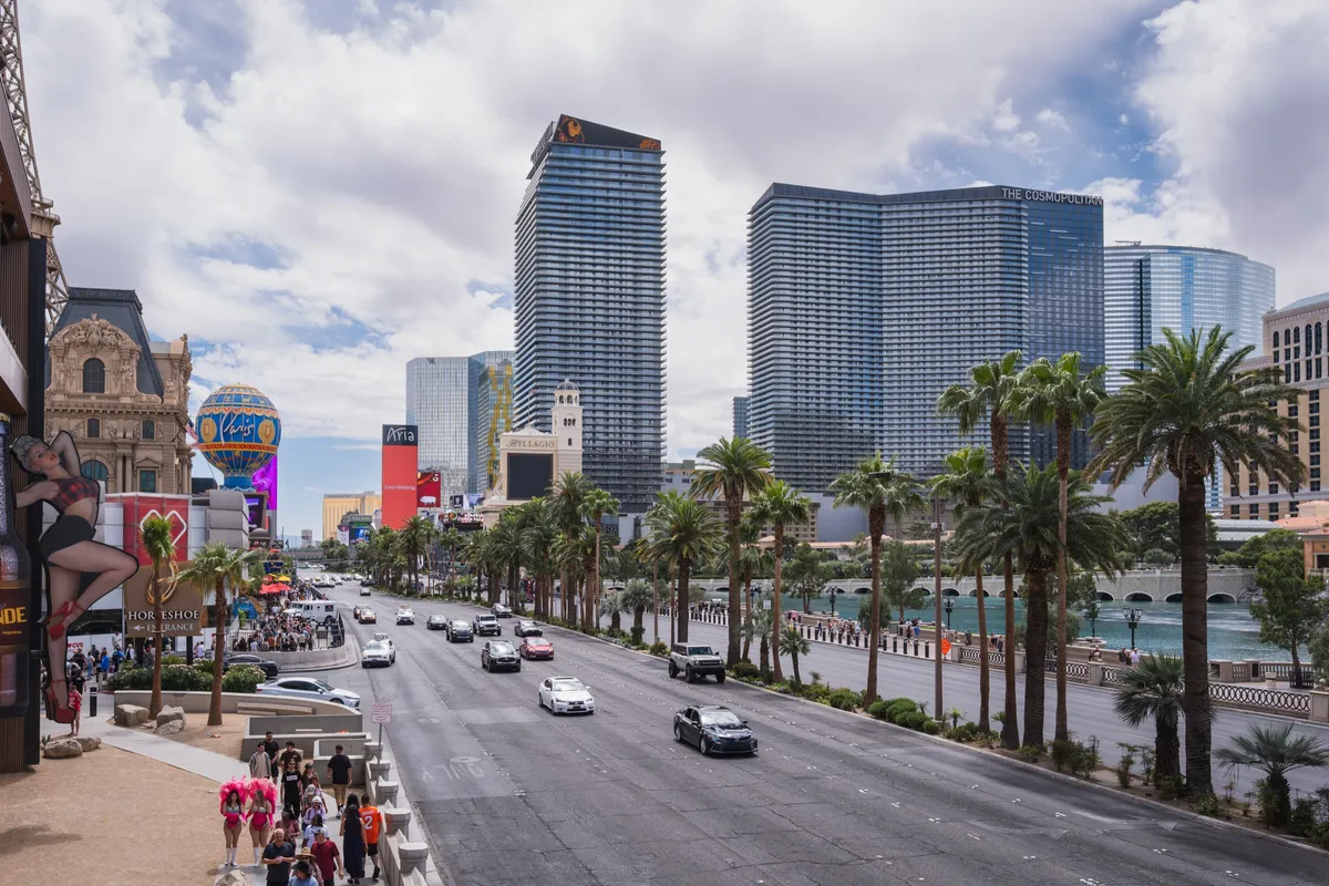 The Las Vegas Strip on a bright day, lined with palm trees and resort towers, with traffic and pedestrians.